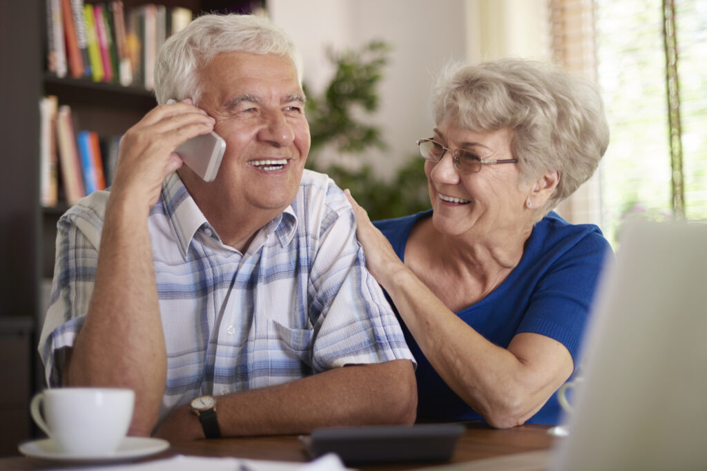 Zu sehen ist ein älteres Ehepaar, Der Mann hat ein Telefon in der Hand. Die Frau lächelt ihn an.